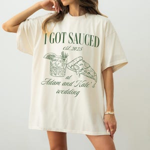 May include: A natural colored t-shirt with a green graphic design that says "I got Sauced est. 2025 at Adam and Kate's wedding". The graphic includes a cocktail glass and a slice of pizza.