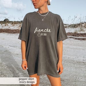 Peut inclure: T-shirt gris fonc&eacute; avec le mot "fianc&eacute;e" en &eacute;criture cursive et "EST. 2026" imprim&eacute; sous un motif d'anneau. Le t-shirt a une coupe ample et des manches courtes. Le texte "pepper shirt ivory design" est en bas.