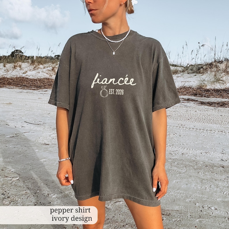 Peut inclure: T-shirt gris fonc&eacute; avec le mot "fianc&eacute;e" en &eacute;criture cursive et "EST. 2026" imprim&eacute; sous un motif d'anneau. Le t-shirt a une coupe ample et des manches courtes. Le texte "pepper shirt ivory design" est en bas.