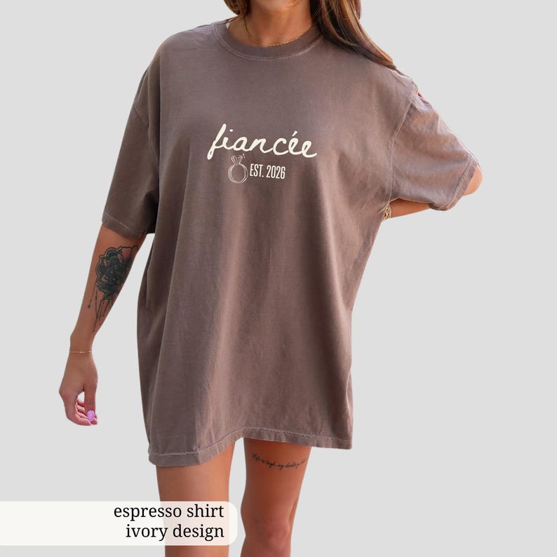 Peut inclure: T-shirt couleur expresso avec le mot "fianc&eacute;e" en &eacute;criture cursive, accompagn&eacute; d'un motif d'anneau et du texte "EST. 2026". Le t-shirt a une coupe ample et des manches courtes. Le design est de couleur ivoire.
