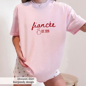 Peut inclure: Un t-shirt rose clair avec le mot "fianc&eacute;e" en &eacute;criture rouge, accompagn&eacute; d'un dessin d'anneau et de "EST. 2026". Le t-shirt a des manches courtes et une coupe ample, id&eacute;al pour un usage quotidien.