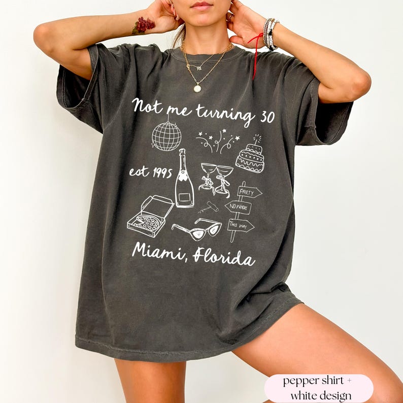 May include: A dark gray oversized t-shirt with a white graphic design that says "Not me turning 30 est 1995 Miami, Florida". The graphic includes a disco ball, champagne bottle, cake, glasses, pizza, sunglasses, and a sign that says "Party No Padre This Way".