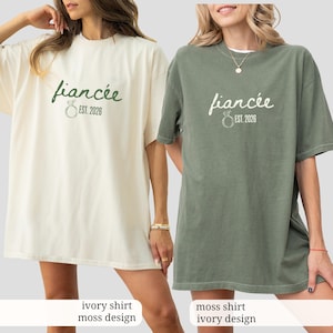 Peut inclure: Deux t-shirts amples, un ivoire et un vert mousse, chacun avec le mot "fianc&eacute;e" en cursive et "EST. 2026" imprim&eacute; sous un motif d'anneau. Le t-shirt ivoire a un motif vert mousse, tandis que le t-shirt mousse a un motif ivoire.