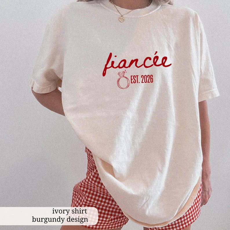 Peut inclure: T-shirt de couleur ivoire avec le mot "fianc&eacute;e" en rouge, un dessin d'anneau et "EST. 2026". Le t-shirt est associ&eacute; &agrave; un short &agrave; carreaux rouges et blancs. L'ensemble est de style d&eacute;contract&eacute;.