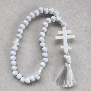 May include: A white prayer rope featuring round beads and two cross-shaped pendants. The rope has a tassel at the end. The beads and crosses are white, and the tassel is a light grey colour. The prayer rope is likely used for religious practices.