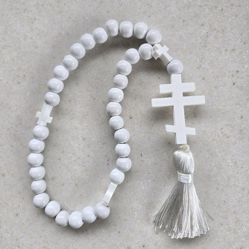 May include: A white prayer rope featuring round beads and two cross-shaped pendants. The rope has a tassel at the end. The beads and crosses are white, and the tassel is a light grey colour. The prayer rope is likely used for religious practices.