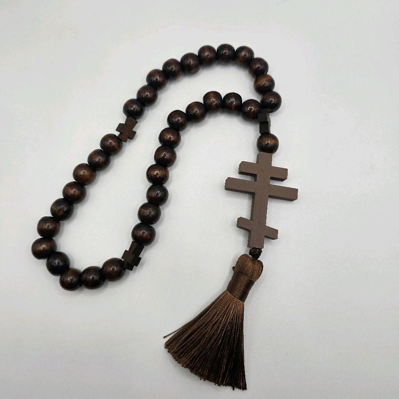 May include: A dark brown wooden prayer rope featuring a series of round beads and two cross pendants. A tassel hangs from the bottom of the larger cross. The beads and crosses are made of wood.