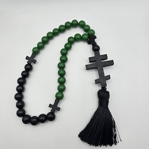 May include: A rosary featuring green and black wooden beads. The rosary has black wooden crosses and a black tassel. The beads are round and evenly spaced, creating a symmetrical design.