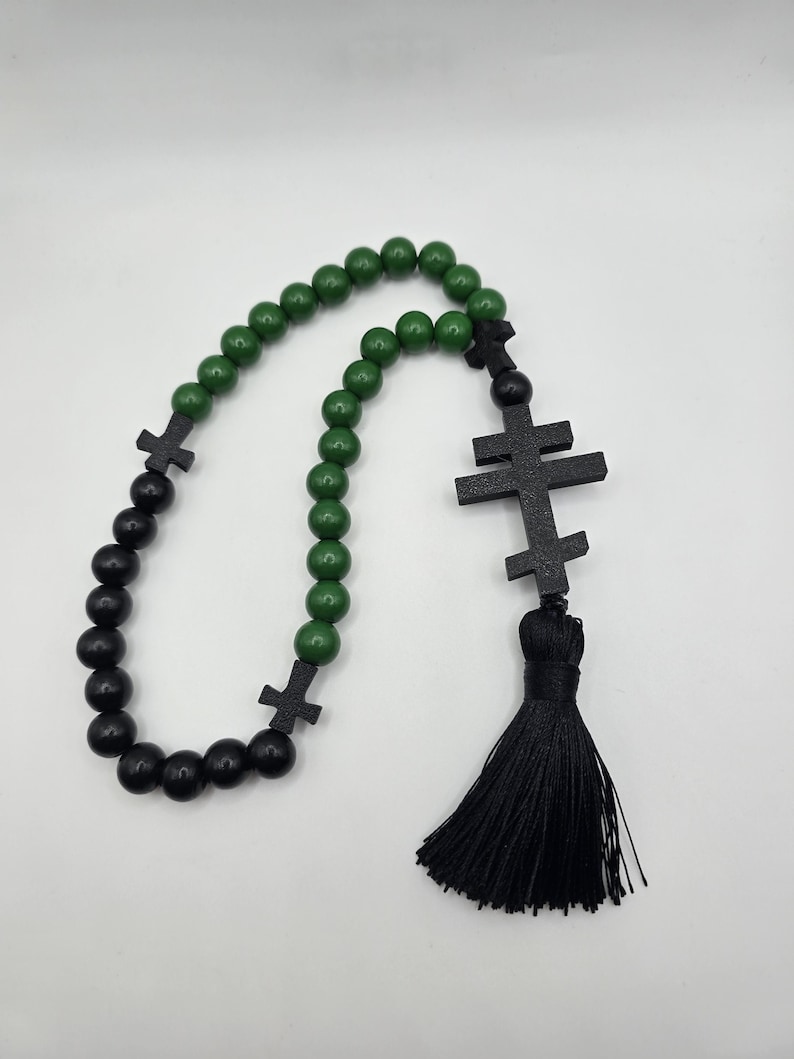 May include: A rosary featuring green and black wooden beads. The rosary has black wooden crosses and a black tassel. The beads are round and evenly spaced, creating a symmetrical design.