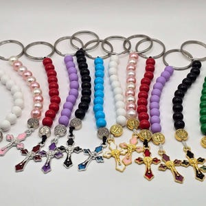 May include: A selection of rosary keyrings in various colours. Each keyring has a string of beads in colours such as white, pink, red, purple, black, blue, and green. The keyrings have a silver cross pendant and a silver keyring.