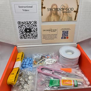 May include: An open orange box containing rosary-making supplies. Includes blue beads, silver-colored charms, a yellow measuring tape, wire, scissors, and glue. The box also contains cards with the text "SWORD OF GOD ROSARIES" and "Instructional Video:" with a QR code.