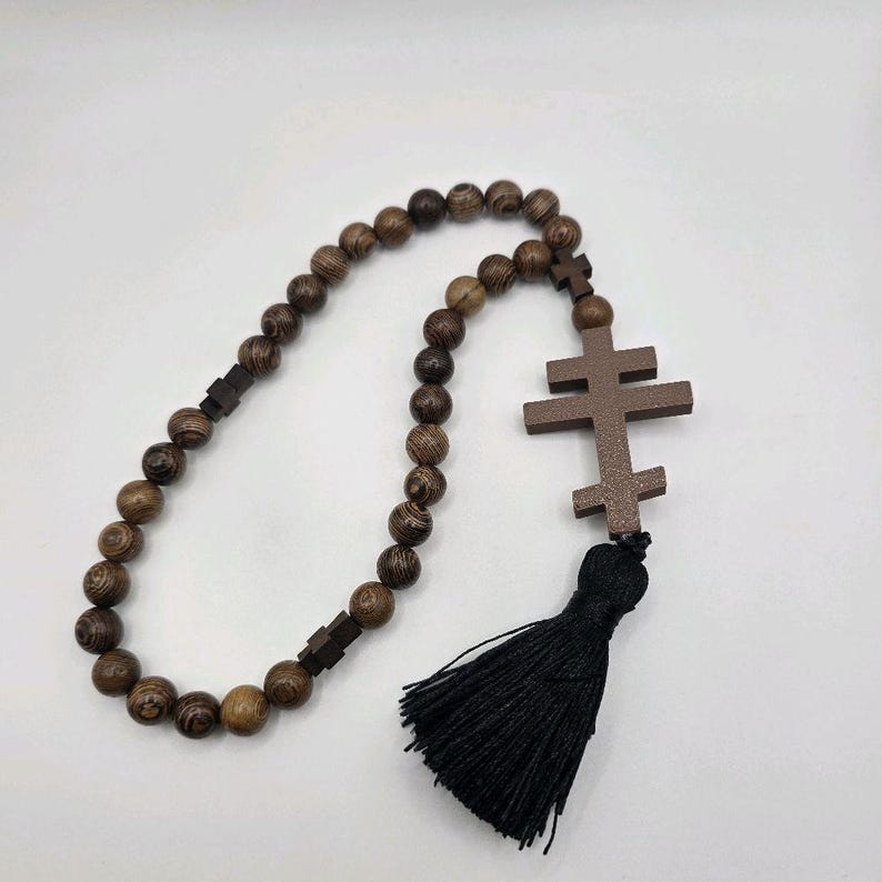 May include: A wooden rosary featuring dark brown beads and a double-barred cross pendant. The rosary has a black tassel. The beads are round and made of wood.