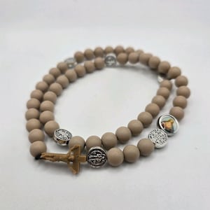 May include: A beige rosary with round beads and silver-colored metal accents. The rosary features a small crucifix and several decorative medallions. The beads are evenly spaced, creating a symmetrical design.
