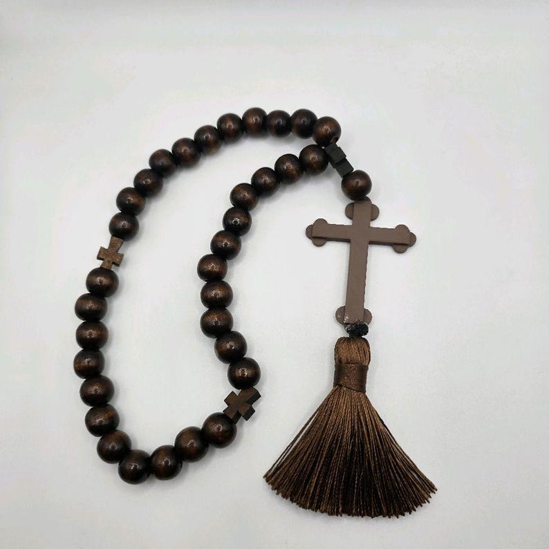 May include: A dark brown rosary with wooden beads and a cross pendant. The rosary features a tassel and small cross accents. The beads are evenly spaced, and the cross is a simple, traditional design. The rosary is likely used for prayer and meditation.