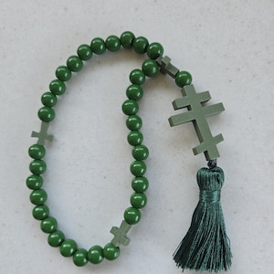 May include: A dark green rosary featuring spherical beads and three cross-shaped elements. A dark green tassel is attached. The rosary is made of wood and is used for prayer and meditation.