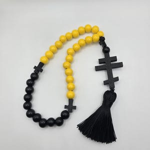 May include: A prayer rope with alternating yellow and black beads. The rope features black cross-shaped accents and a black tassel. The beads are round and evenly spaced, creating a symmetrical design. The background is plain white.