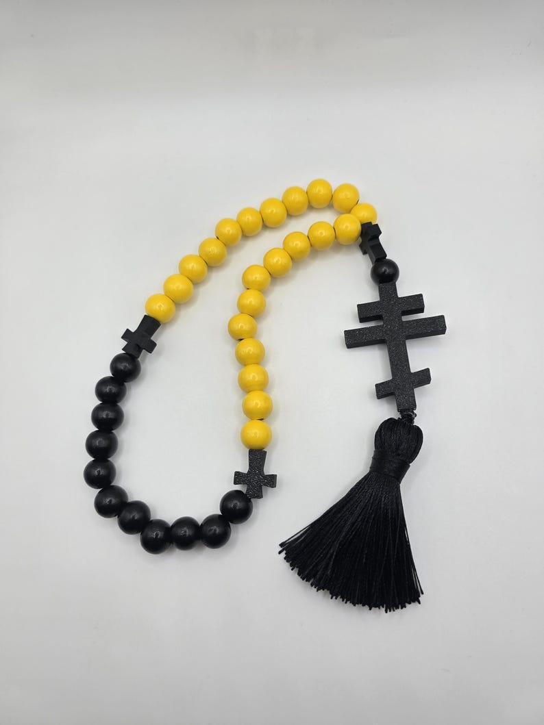 May include: A prayer rope with alternating yellow and black beads. The rope features black cross-shaped accents and a black tassel. The beads are round and evenly spaced, creating a symmetrical design. The background is plain white.