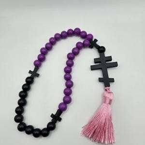 May include: A rosary featuring purple and black beads, black cross-shaped dividers, and a pink tassel. The beads are arranged in a loop. The tassel hangs at the bottom. The background is plain white.
