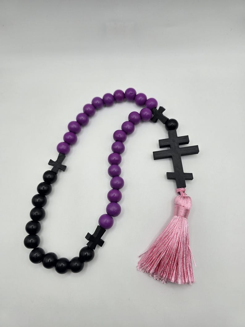 May include: A rosary featuring purple and black beads, black cross-shaped dividers, and a pink tassel. The beads are arranged in a loop. The tassel hangs at the bottom. The background is plain white.