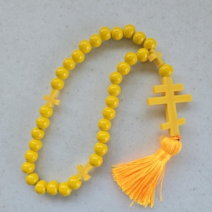 May include: A yellow beaded rosary featuring a double-barred cross pendant and two smaller crosses. It has a bright orange tassel. The beads are round and evenly spaced.