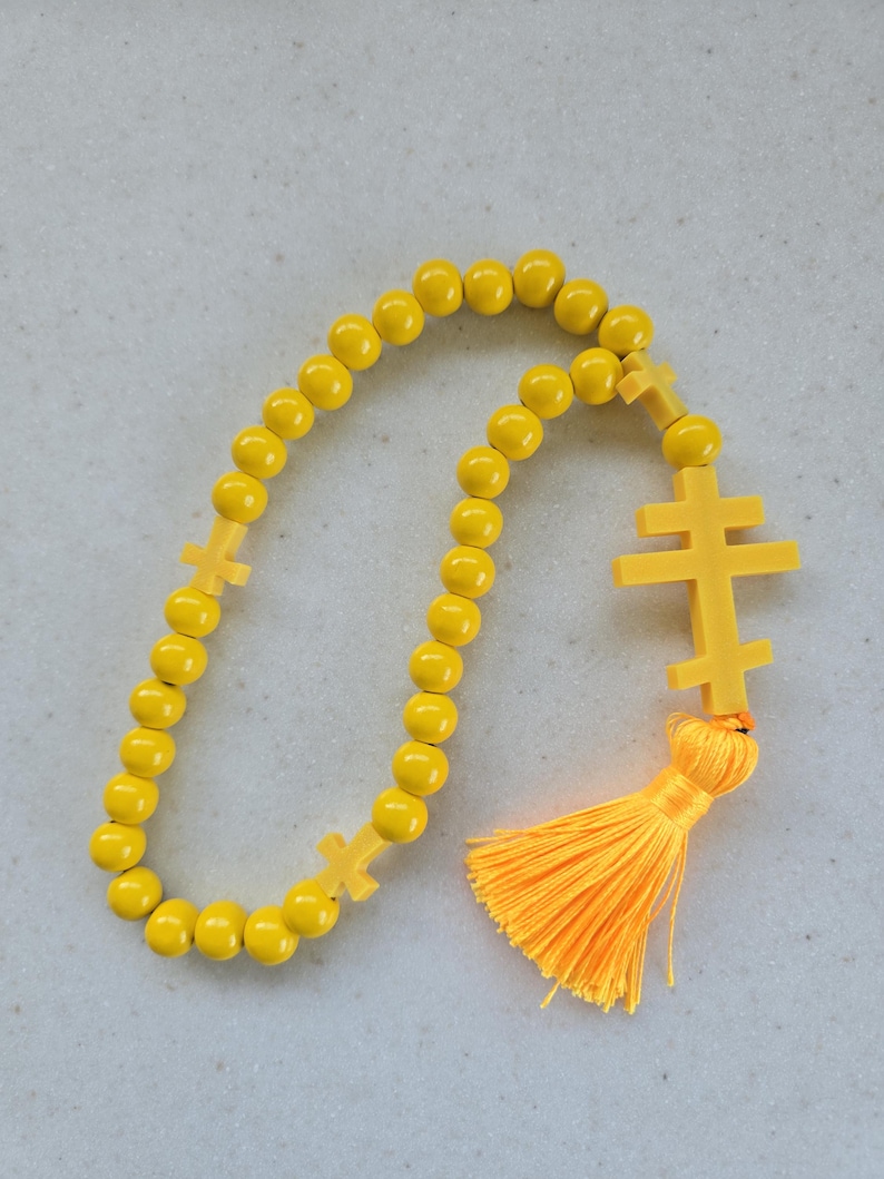 May include: A yellow beaded rosary featuring a double-barred cross pendant and two smaller crosses. It has a bright orange tassel. The beads are round and evenly spaced.