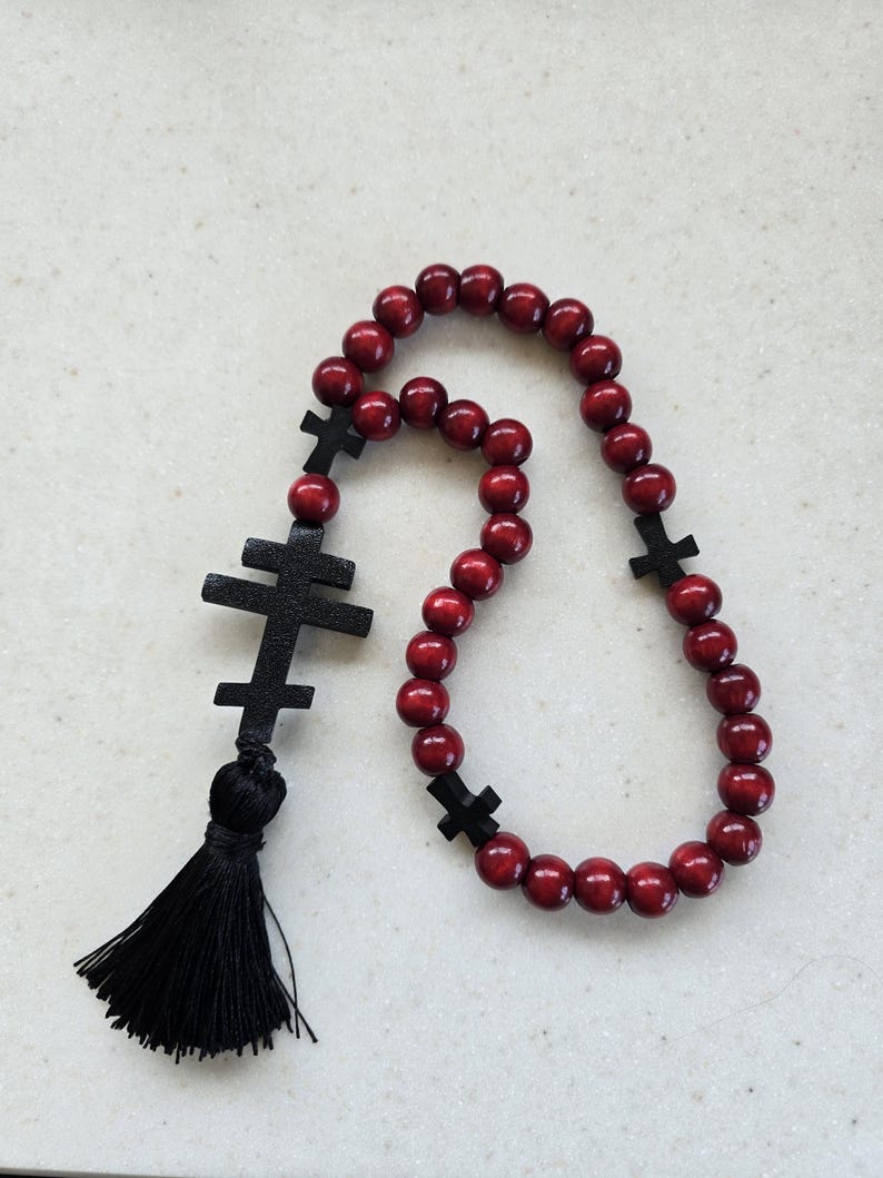 May include: A red wooden prayer rope featuring black crosses and a black tassel. The prayer beads are round and evenly spaced. The crosses are of varying sizes and shapes, with the largest at the top.