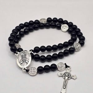Puede incluir: Rosario de cuentas negras con crucifijo plateado y colgante del Sagrado Corazón de Jesús. El rosario presenta cuentas negras redondas y varias medallas plateadas. El crucifijo tiene las letras INRI.