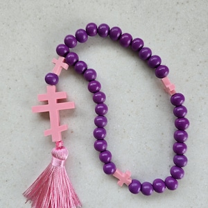 May include: A purple beaded rosary with pink cross accents and a pink tassel. The rosary features round, dark purple beads and three pink crosses. A pink tassel hangs from the end of the rosary.