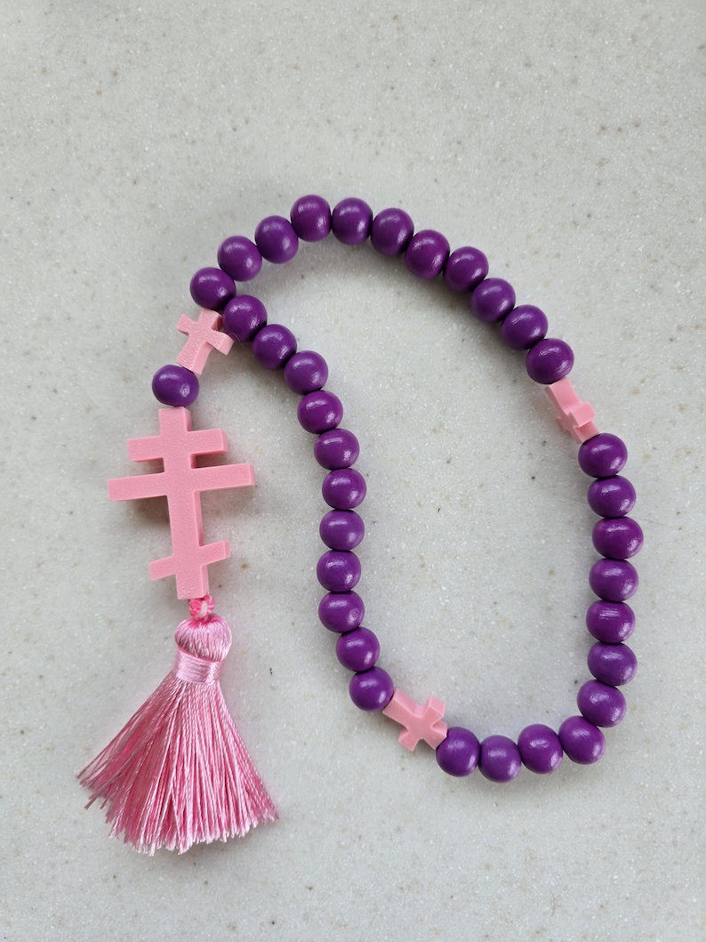 May include: A purple beaded rosary with pink cross accents and a pink tassel. The rosary features round, dark purple beads and three pink crosses. A pink tassel hangs from the end of the rosary.