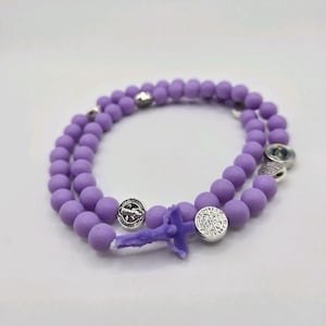May include: A rosary bracelet featuring matte lavender beads arranged in a circular pattern. The bracelet includes several small, round silver-toned metal charms with embossed religious imagery and a small purple crucifix pendant attached.