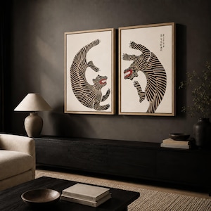 May include: Two rectangular framed wall art pieces featuring stylized black and white tiger illustrations on a cream background. Each tiger is depicted in a dynamic, curved pose with visible red accents in the mouth area and black calligraphy text.