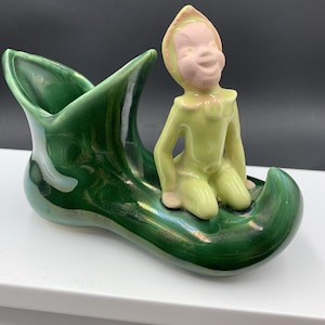 Könnte beinhalten: Vintage Keramikfigur mit einer grünen stiefelförmigen Pflanzschale und einer sitzenden Elfenfigur. Der Elf hat ein rosa Gesicht und ein gelb-grünes Outfit. Der Stiefel hat eine glänzende Oberfläche mit schillernden Highlights, was ein skurriles Dekorationsobjekt ergibt.