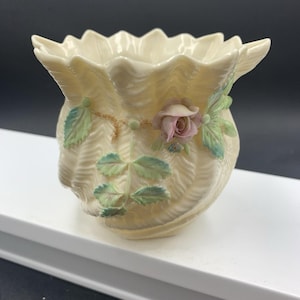 Puede incluir: Un jarrón de porcelana color crema con un borde festoneado similar a una concha y un cuerpo texturizado y acanalado. El jarrón está decorado con delicados detalles de cerámica tridimensionales que presentan una rosa rosa y hojas verdes en una enredadera.
