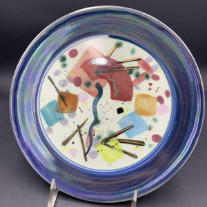 May include: A ceramic plate with a blue rim and a white center. The center features colorful abstract shapes in red, orange, blue, and yellow, with brown and black accents. The plate is displayed on a silver stand.