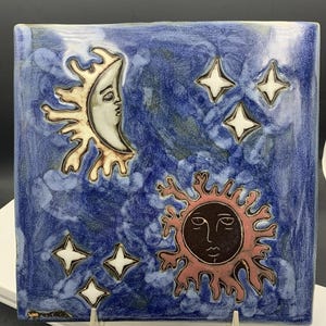 May include: A square ceramic tile featuring a deep blue glazed background with a hand-painted celestial scene. The design includes a cream-colored crescent moon with a face, a dark red sun with a face, and five white four-pointed stars.