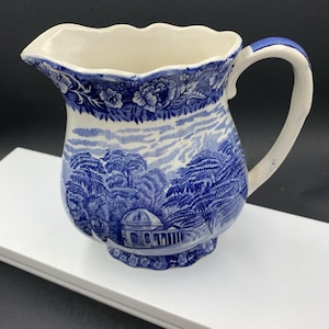 May include: A blue and white ceramic pitcher with a landscape design. The pitcher has a scalloped rim and a curved handle. The design features trees, buildings, and floral patterns. The pitcher is approximately 6 inches tall.