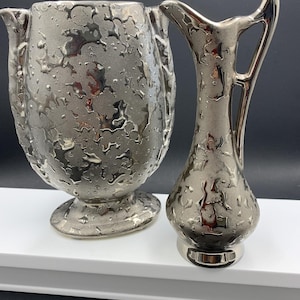 Könnte beinhalten: Zwei silberfarbene dekorative Vasen. Die größere Vase hat eine bauchige Form mit strukturierter Oberfläche und zwei Ausgüssen. Die kleinere Vase ist ein schlanker Krug mit Henkel und ähnlicher strukturierter Oberfläche. Beide stehen auf einer weißen Oberfläche.