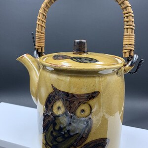 Könnte beinhalten: Keramik-Teekanne mit beigefarbener Glasur und Eulen-Design. Sie hat einen geflochtenen Rattangriff und einen Deckel mit kleinem Knopf. Die Eule ist in Braun- und Grüntönen dargestellt. Ein dekorativer Küchenartikel.