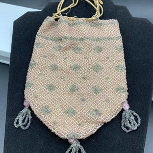 Può includere: Un'antica borsetta a coulisse di perline in una delicata tonalità rosa, ornata da un delicato motivo di piccoli motivi floreali grigio-verdi. La borsa presenta nappe di perline sul fondo e una chiusura a coulisse intrecciata.