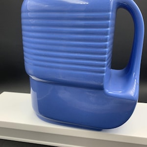 May include: A vintage blue ceramic jug with a ribbed design on the upper section. The jug has a curved handle and a spout for pouring. The jug is on a white surface against a black background. Suitable for kitchen decor, this item is a classic.
