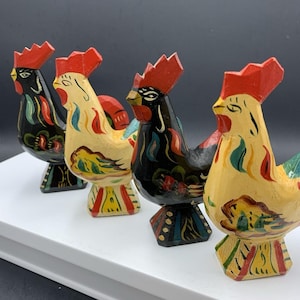 May include: Four hand-painted wooden cockerel figurines arranged in a row. Two cockerels are painted black with red combs and colourful floral accents, while the other two are painted yellow with red combs and similar multi-coloured decorative patterns.