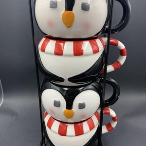 Pode incluir: Um conjunto de três canecas em forma de pinguim empilhadas em um suporte de metal preto. As canecas são brancas com detalhes em preto, cachecóis listrados em vermelho e branco e bicos laranja. Cada caneca tem uma alça preta.