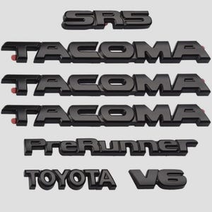 Può includere: Emblemi automobilistici neri tra cui "SRS", più badge "Tacoma", "PreRunner", "Toyota" e scritte "V6". Gli emblemi sono in un font sans-serif e hanno una finitura lucida.