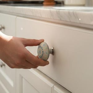 May include: A hand opening a white kitchen cabinet drawer with a decorative ceramic knob. The knob has a light blue and white floral design. Other cabinets have silver knobs. A small potted plant sits on the countertop.