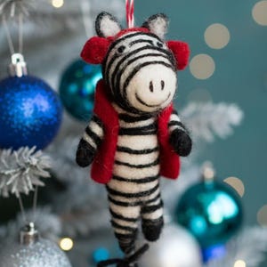 May include: A handmade Christmas ornament shaped like a zebra. The zebra has black and white stripes, red ears and a red vest. The ornament is hanging from a white and red string, with a Christmas tree and ornaments in the background.