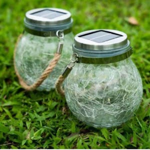 May include: Two solar-powered jar lanterns with a crackled glass design. Each lantern has a silver lid with a solar panel, a metal band, and a rope handle. The lanterns are on green grass.