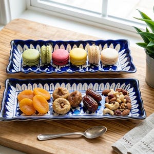 May include: Two rectangular blue and white patterned serving dishes. One holds colorful macarons, the other dried apricots, figs, dates, and nuts. A small potted plant and a spoon are also present.