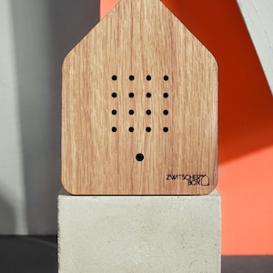 Puede incluir: Un objeto de madera con forma de casa con una serie de pequeños agujeros, apoyado sobre un bloque de hormigón. La veta de la madera es visible. El texto "ZWITSCHER BOX" está impreso en la parte inferior derecha.