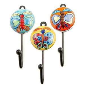 Pode incluir: Três ganchos de metal preto com botões de cerâmica de borboleta coloridos. As borboletas são pintadas em tons de vermelho, azul, amarelo e verde.