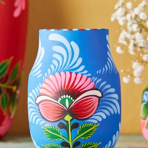 Può includere: Un vaso blu con un motivo floreale dipinto a mano. Il vaso presenta un grande fiore rosa e rosso con foglie verdi e dettagli bianchi. È un oggetto decorativo.